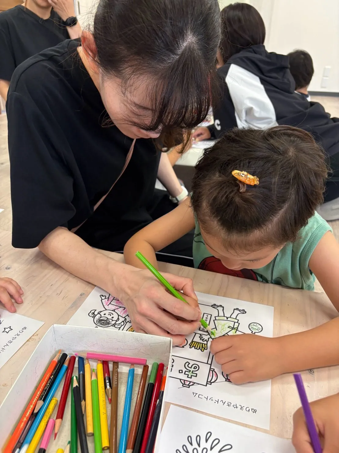 机で子どもの隣に座り、色鉛筆で塗り絵に取り組む様子を見守るスタッフ