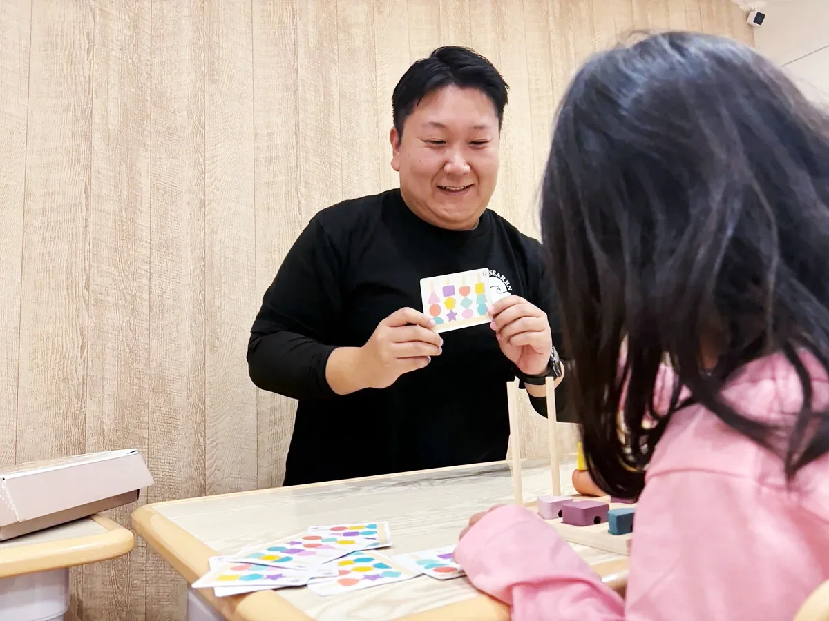 木のおもちゃが置かれた机で、子どもにカードを見せながら関わるスタッフ